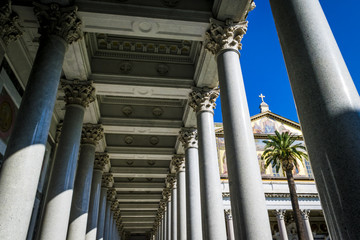 Marble columns and ornate ceiling of St. Paul’s Cathedral in Rome, Italy.  Architectural detail, blue sky, facade of the Cathedral and a palm tree in background.