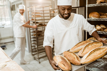 handsome african american baker taking bread loaves from oven at baking manufacture