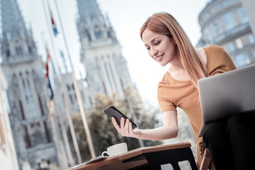 Reading news. Low angle shot of a positive minded woman browsing the Internet on her phone and smiling cheerfully while sitting outdoors.