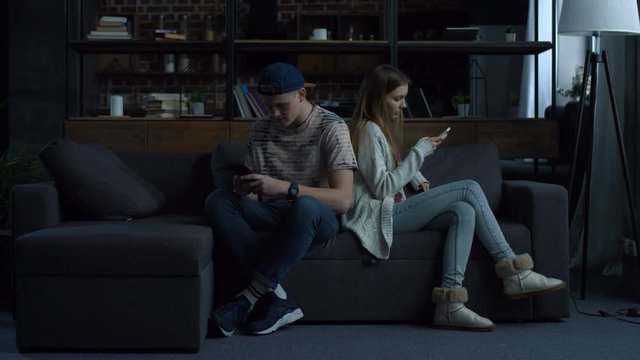 Attractive young couple sitting back to back at home on couch obsessed with smartphones. Couple with mobile phones ignoring each other as strangers, communication problems and social network addiction