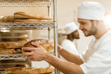 handsome smiling  baker putting fresh bread loaf on shelf at baking manufacture