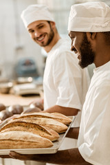 handsome young bakers in hats at baking manufacture looking at fresh bread loaves on tray