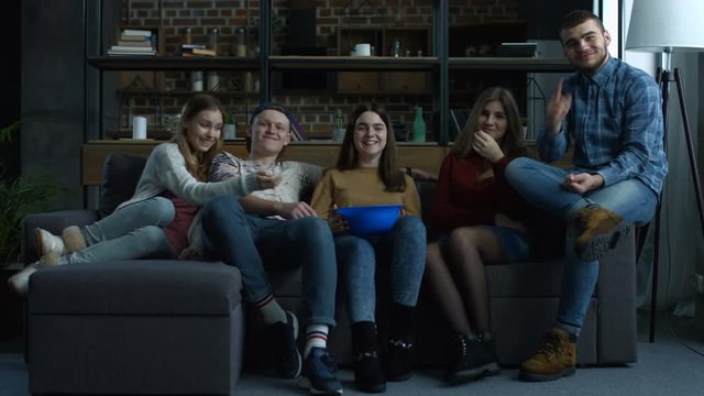 Group of cheerful teenage friends watching comedy movie on TV with popcorn and laughing while sitting on sofa at home. Excited casual friends relaxing and watching television in living room.