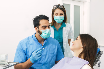 Dentist checking teeth, young asian man, in blue uniform and gloves, looking at patient's teeth carefully with mouth mirror and explorer, pretty caucasian brunette sitting patiently in dental chair