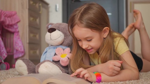 Cute happy little girl with teddy bear and reading book. Pretty kid at home, lying on the floor. Education concept