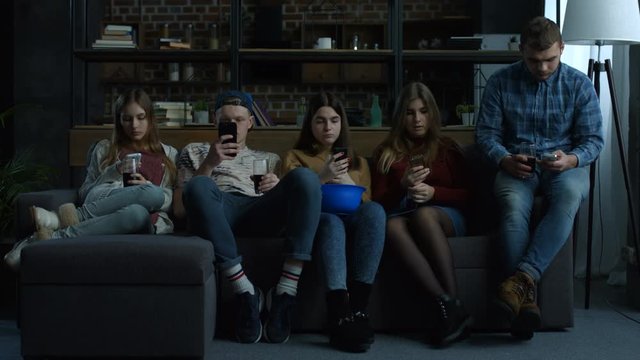 Group of young friends phubbing each other while sitting in a row on the couch in domestic room. Busy teenagers using smart phones with disinterest on each other. Technology and smart phone addiction.