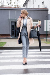 businesswoman with coffee to go and umbrella talking on smartphone while crossing road on street