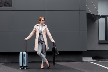 stylish businesswoman in coat with suitcase waiting for taxi on street