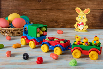 Obraz premium Easter toy car with trailer and color sweets in, Easter eggs, candy and bunny on wooden background