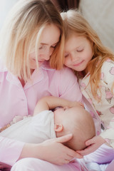 mom blonde with older daughter look at the newborn baby sister and smile 