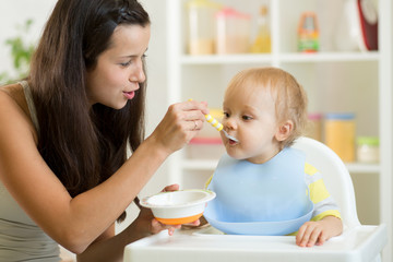 Mom feeding hungry baby son in the highchair indoors