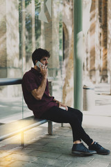 Side view portrait of young hi[ster guy chatting happily with his friend by a mobile phone. Handsome model look guy talking on a phone while waiting for a bus on a sunday morning.