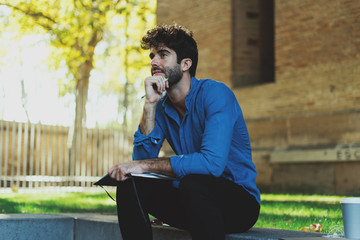 Obraz premium International student preparing for the exams while sitting in a park. Thoughtful bearded man in a light blue shirt sitting with a pencil and a notebook in his hands with a coffee standing beside.