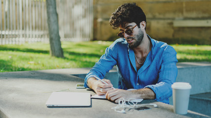 Portrait of a model look caucasian male wearing trendy sunglasses sitting at the table with a laptop and take away coffee cup on it. Office worker preparing for the meeting during coffee break.