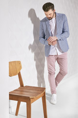 Fashion portrait of handsome male model with dark hair and beard wearing light pants, white shirt, plaid jacket with chair near him