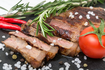 Sliced roasted prime Black Angus beef steak strip loin with rosemary herb, salt, tomatoes and red chili peppers on a black board.