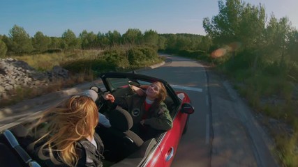 Two best friends or girlfriends make selfie video or self portrait photo on stick action camera, while stand up inside convertible cabriolet car on mountain road. Happiness friendship and freedom
