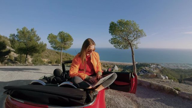 Steadycam shot of young natural beauty attractive girl or woman, new generation always connected millennial work remotely from office on laptop, in red convertible cabriolet outdoors in nature