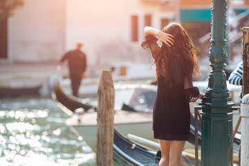 Fototapeta premium Travel tourist girl on vacation walking happy by Grand Canal. Attractive young romantic woman standing on the pier against beautiful view on venetian chanal with boats and gondolas in Venice, Italy