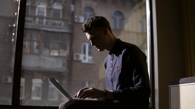 Attractive sexy young guy sitting near panoramic window typing something on his laptop. Work process.