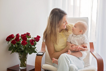 Young mother holding her baby boy, playing with him, sitting in armchair