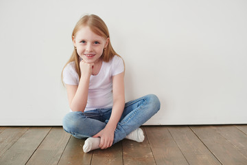 Portrait of a little girl sitting on a wooden floor near a white wall