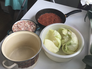 On the table are the ingredients for cooking cabbage rolls. Puffed carrots in a frying pan, minced meat in a container and cabbage leaves in a basin.