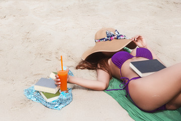 The  lady  is lay down on the beach and cover her face with woven hat,she is holding glass of orange juice with right hand and put the book in her body,on the beach,blurry light around.