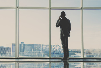 Back view of young businessman is looking out of a panoramic window and talking by phone