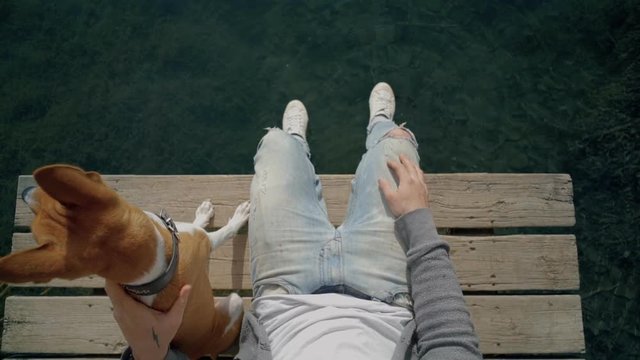 Pov shot of man in blue light denim jeans and white sneakers sit on edge of boardwalk on lake or ocean with best friend puppy dog, makes ripples on water with his shoe, concept nature lifestyle