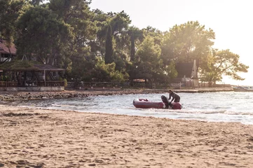 Foto op Canvas Man duwt zijn opblaasbare boot naar het strand in de buurt van een openbare plaats met bomen © Ipek Morel