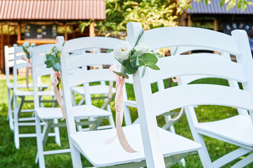 White fresh flowers with pink ribbon on wedding chair on each side of archway outroods; copy space. Wooden chairs for guests on green grass in the garden on wedding ceremony