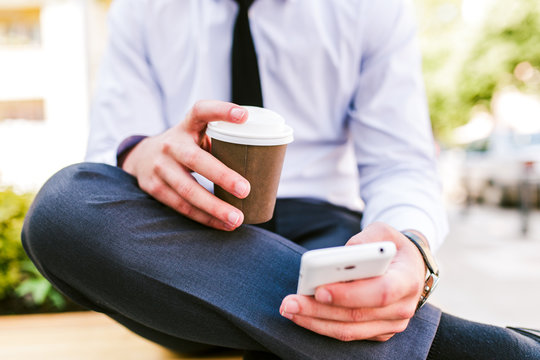 Young businessman using cellphone while sitting outdoor and drinking coffee 