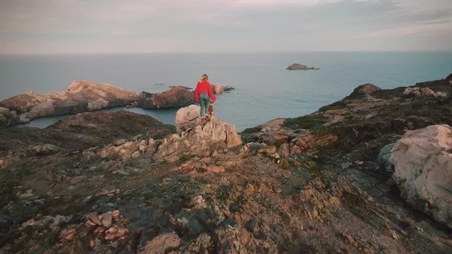 Epic and inspiring drone footage of young happy and excited woman adventurer with best friend dog pet stand on top of mountain in amazing scenery landscape.