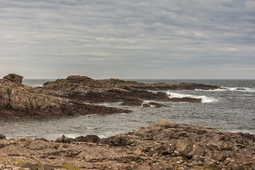 Fototapeta na wymiar Cove, Scotland - June 9, 2012: End-of-road lookout over Atlantic Ocean showing brown rock formation descending in gray water with surf under cloudy sky.