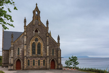 Fototapeta premium Gairloch, Scotland - June 10, 2012: The Free Church of Scotland brown stone building on the shore of the blue-gray Atlantic Ocean. Light blue sky. Vegatation adds green.