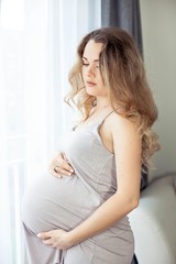 A young beautiful pregnant woman is standing by the window. Pregnant in a beautiful dress. Motherhood. Pregnancy. Portrait of a happy pregnant woman. Cozy.