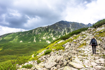 Hiker woman on mountain trail hiking with backpack, path with stones in high mountains, panoramic landscape