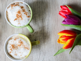 Tulips on wooden background with two cups of coffee. Invitation postcard for mother's day or international women's day. Punchy pastels. Cappuccino with foam cinnamon and flowers.