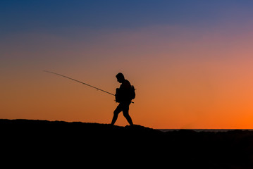 3 Fishermen silhouette with upright rods stroll walk on the rocks at dawn dusk sunrise sunset. The sky is blue purple orange.  They have upright rodsat dawn  