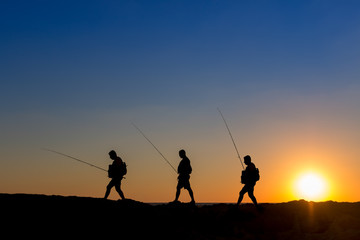 Fototapeta na wymiar 3 Fishermen silhouette with upright rods stroll walk on the rocks at dawn dusk sunrise sunset. The sky is blue purple orange. They have upright rodsat dawn 