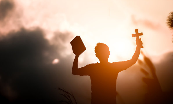 Young man hands holding holy Bible and lift of christian cross with light sunset background. christian concept