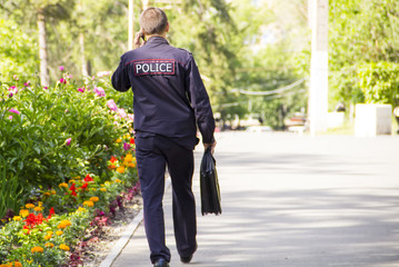 A policeman in uniform with a bag goes to work through the park and talks on his mobile phone.