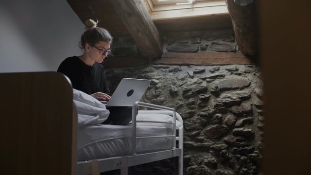 Young independent girl isits on top bunk of two level bed and works on her laptop. She runs her blog or write a code, doing her freelance job at hostel shared room while traveling .