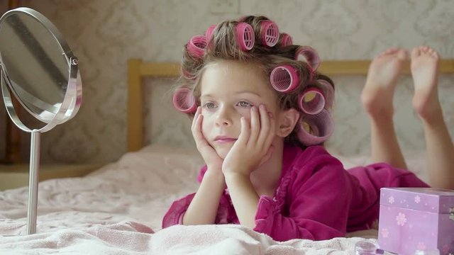 Small girl with rolled hair curlers lies on her bed looking in a mirror