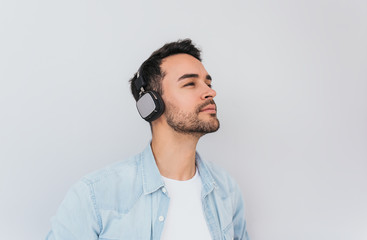Portrait of serious handsome Caucasian male listens music or audio book, uses modern wireless headphones, poses against grey background with copy space for your information advertising or text.