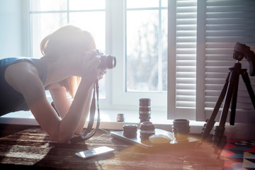 Mid adult woman, beside window, looking through camera, camera lenses on table in front of her