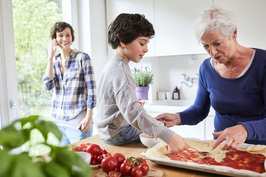 Grandmother and grandson making pizza in kitchen, mother in background using smartphone