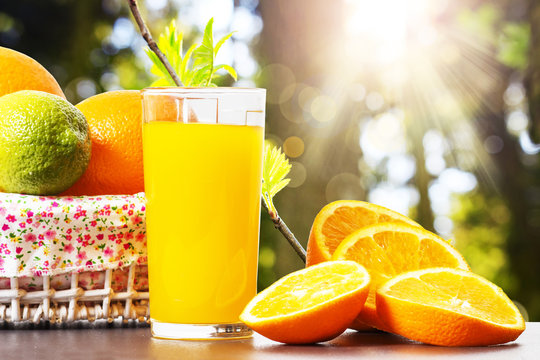 Fresh orange juice in glass beaker, slices of sliced oranges and basket on background of spring green nature with sunbeams. Vitamins and fruits. Healthy lifestyle. Spring background with orange juice.