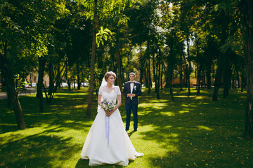 Beautiful wedding photosession. Handsome groom in blue formal suit and his elegant bride in white dress and veil with bouquet with beautiful hairdress on a walk in the big green park on a sunny day